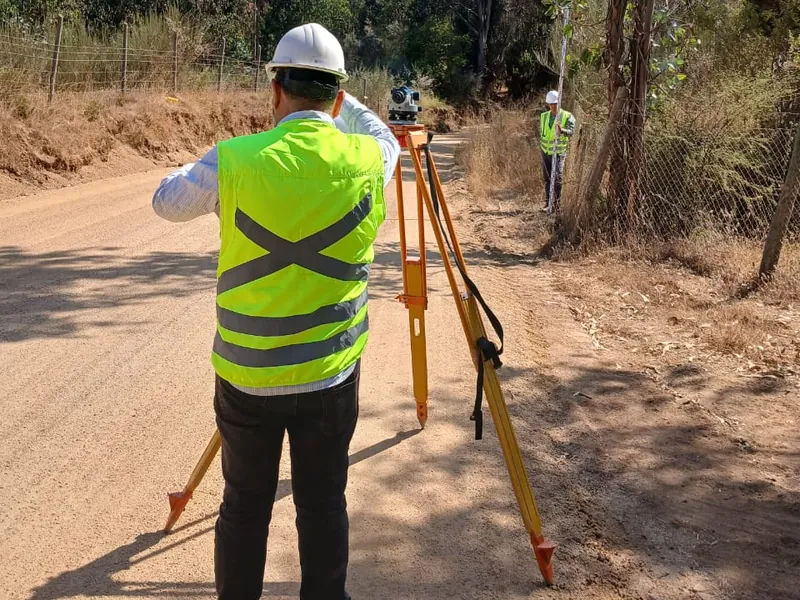 Topógrafo con casco y chaleco reflectante operando un instrumento topográfico sobre trípode en un camino rural, con un jalonero al fondo